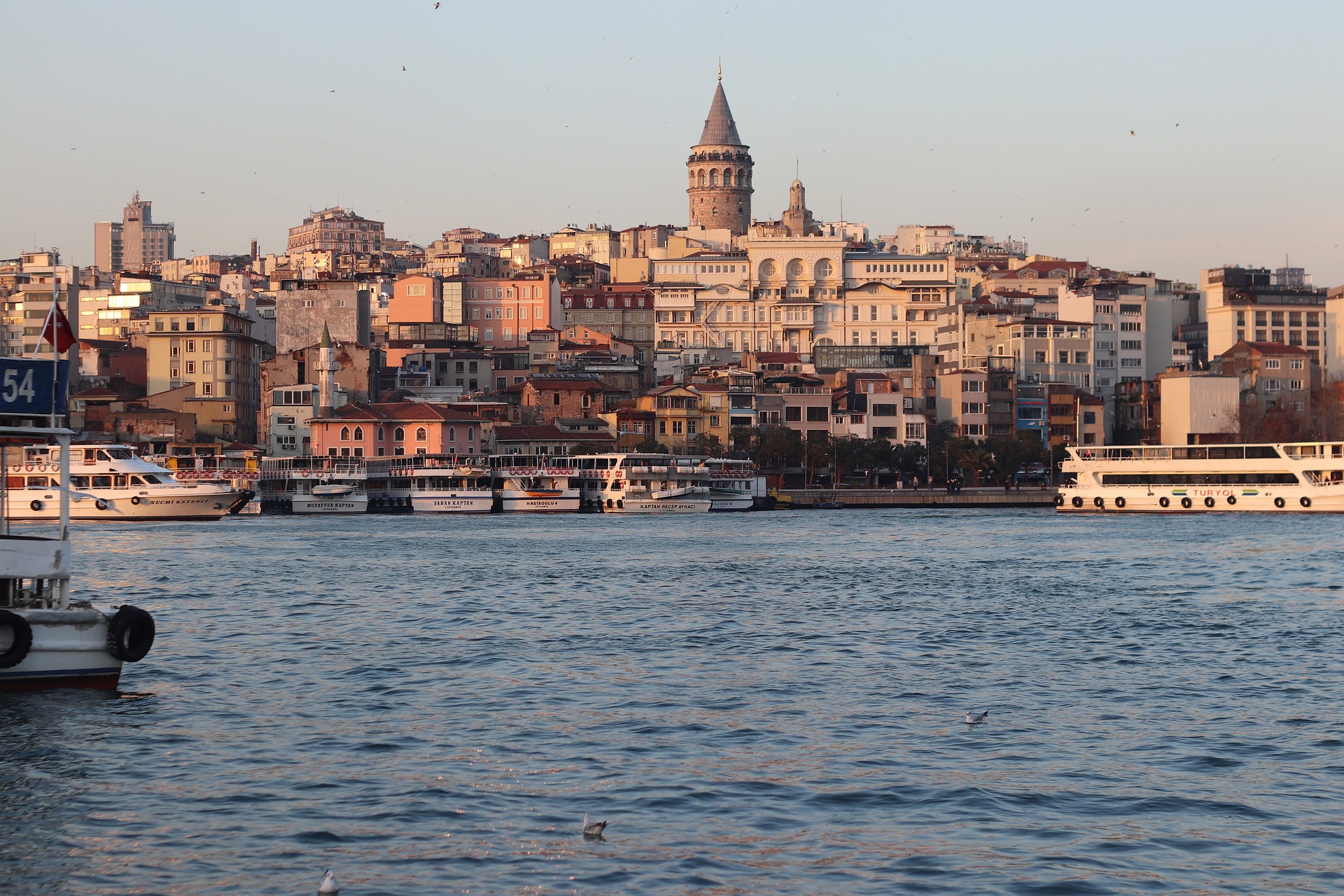 View of the Galata Tower and surrounding buildings along the Bosphorus in Istanbul at sunset, with boats docked by the waterfront.